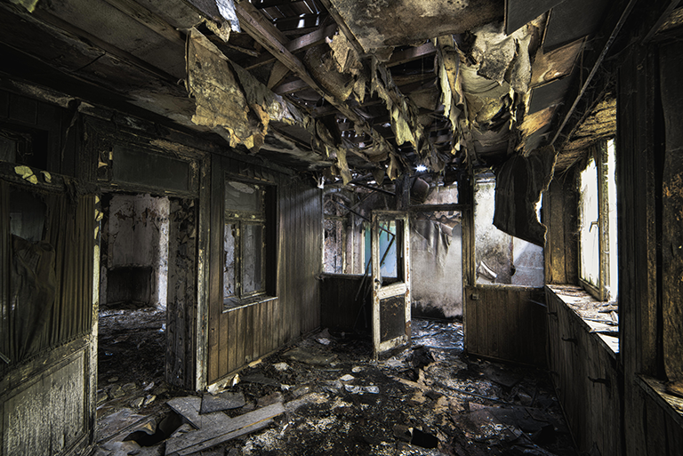 Interior of a severely fire-damaged home with charred walls, collapsed ceiling materials, and debris covering the floor.
