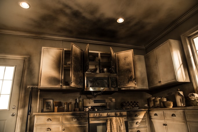Kitchen interior with dry smoke damage, showing soot-stained cabinets and dark residue across the ceiling above the stove.
